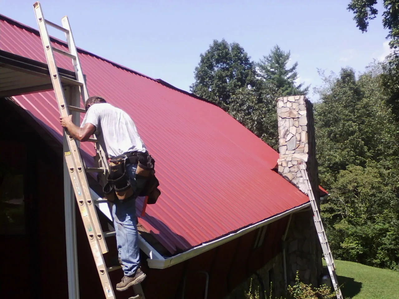 Roofer on a ladder beside a red metal roof and stone chimney — Soffit & Fascia Repair by Roofing Brothers in Masury